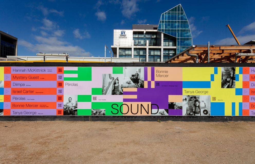 A mural featuring images of musicians and blocks of colours covers construction hoarding in a plaza.