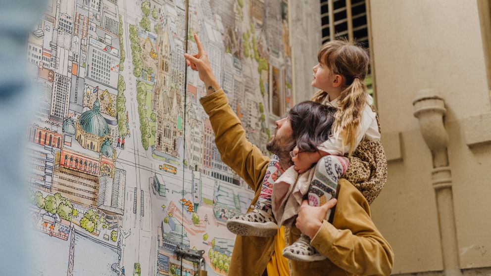A man holds a young girl on his shoulders, who is pointing at the Melbourne Map artwork