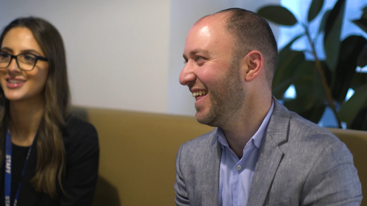 A photo of two smiling people, they are dressed in semi-formal attire and sat in an office setting