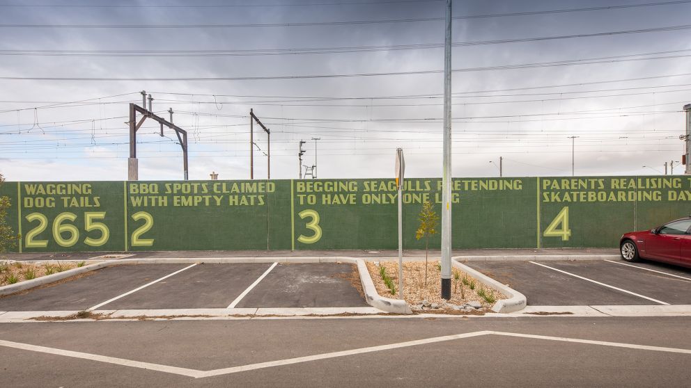 Green and yellow artwork with numbers written on it covers a hoarding wall near a car park