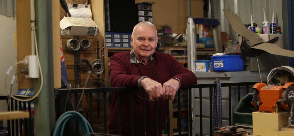 An older man leaning on a railing in a workshop surrounded by tools.