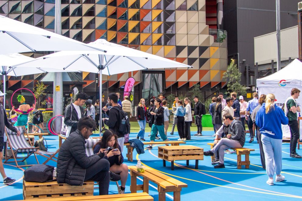 Image of a crowd of people on a blue basketball court. There are white umbrellas and wooden crate seating. 