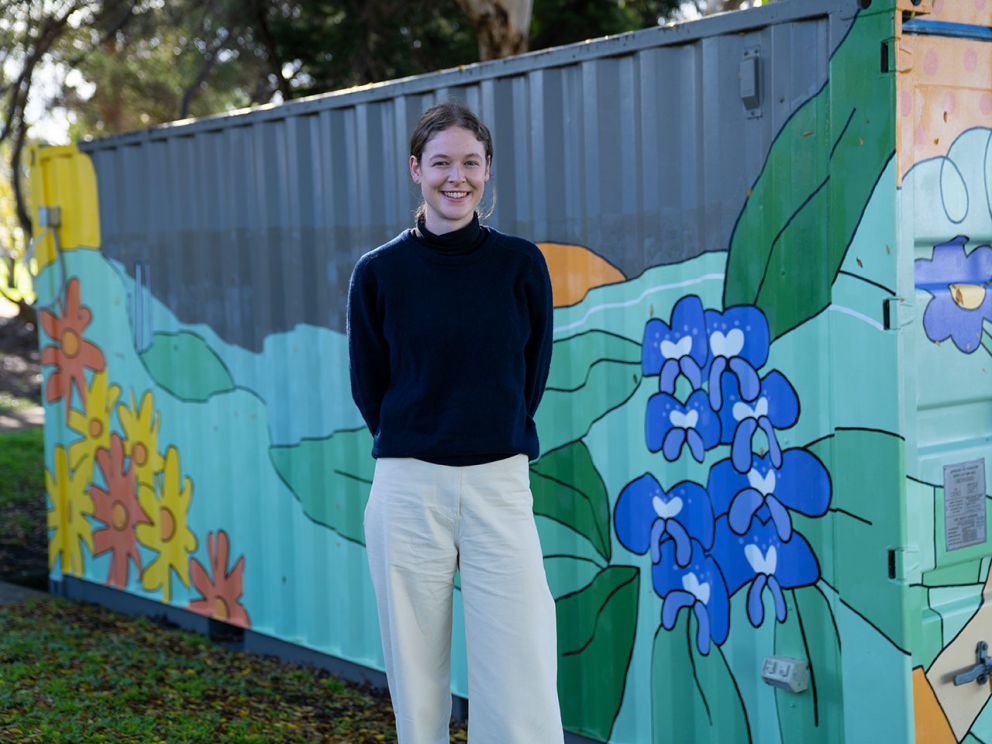 Image shows Melbourne artist Angharad Neal-Williams standing infront of a container painted in bright floral colours. 