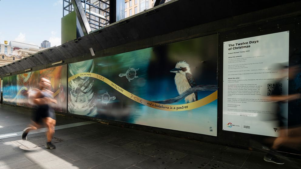 Two people running past colourful construction hoarding that features two tiny turtles and a kookaburra in a gum tree