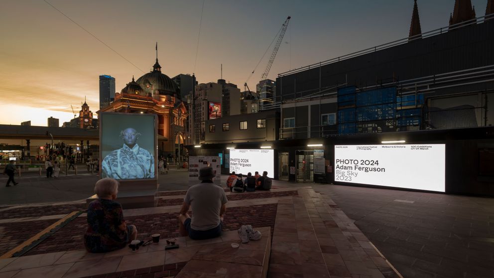 2 people sitting infront of digital screens, in front of a construction site and Flinders Street Station in the background at sunset