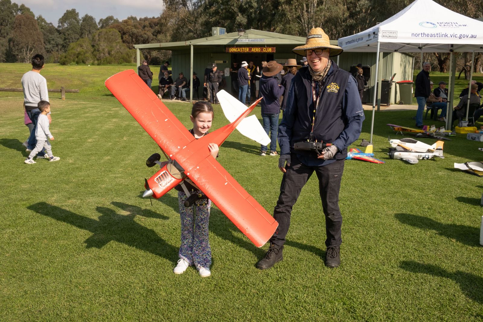 A young girl proudly holds a large red model airplane, standing next to a man wearing a straw hat and glasses, holding a remote control. Both are smiling on a grassy field at what appears to be a model. aircraft event. In the background, people gather near a small green clubhouse 