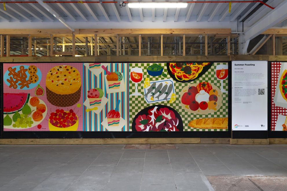 An artwork featuring an assortment of colourful foods on patterned tablecloths installed on a construction hoarding