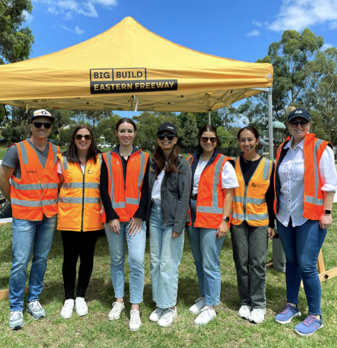 A group of seven people wearing orange safety vests and casual attire stand together in front of a yellow tent labeled "BIG BUILD EASTERN FREEWAY," with grass and trees in the background.