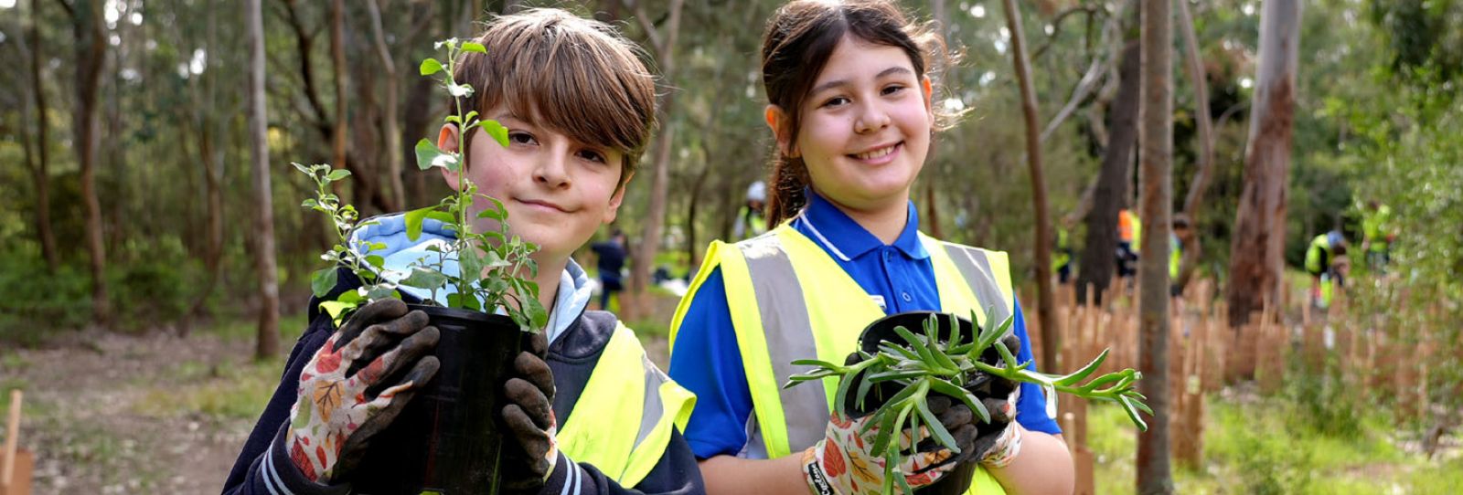 2 kids holding a pot plant each and wearing bright yellow vests. The background features large trees.