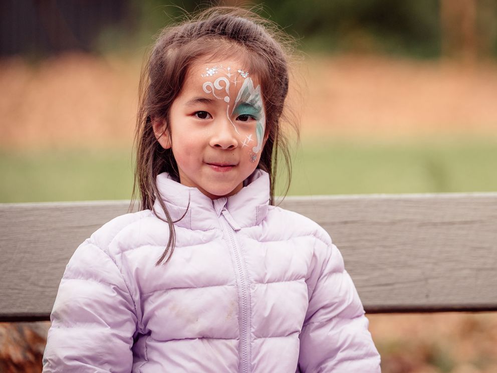 Child with face painted smiling at camera while sitting on a bench