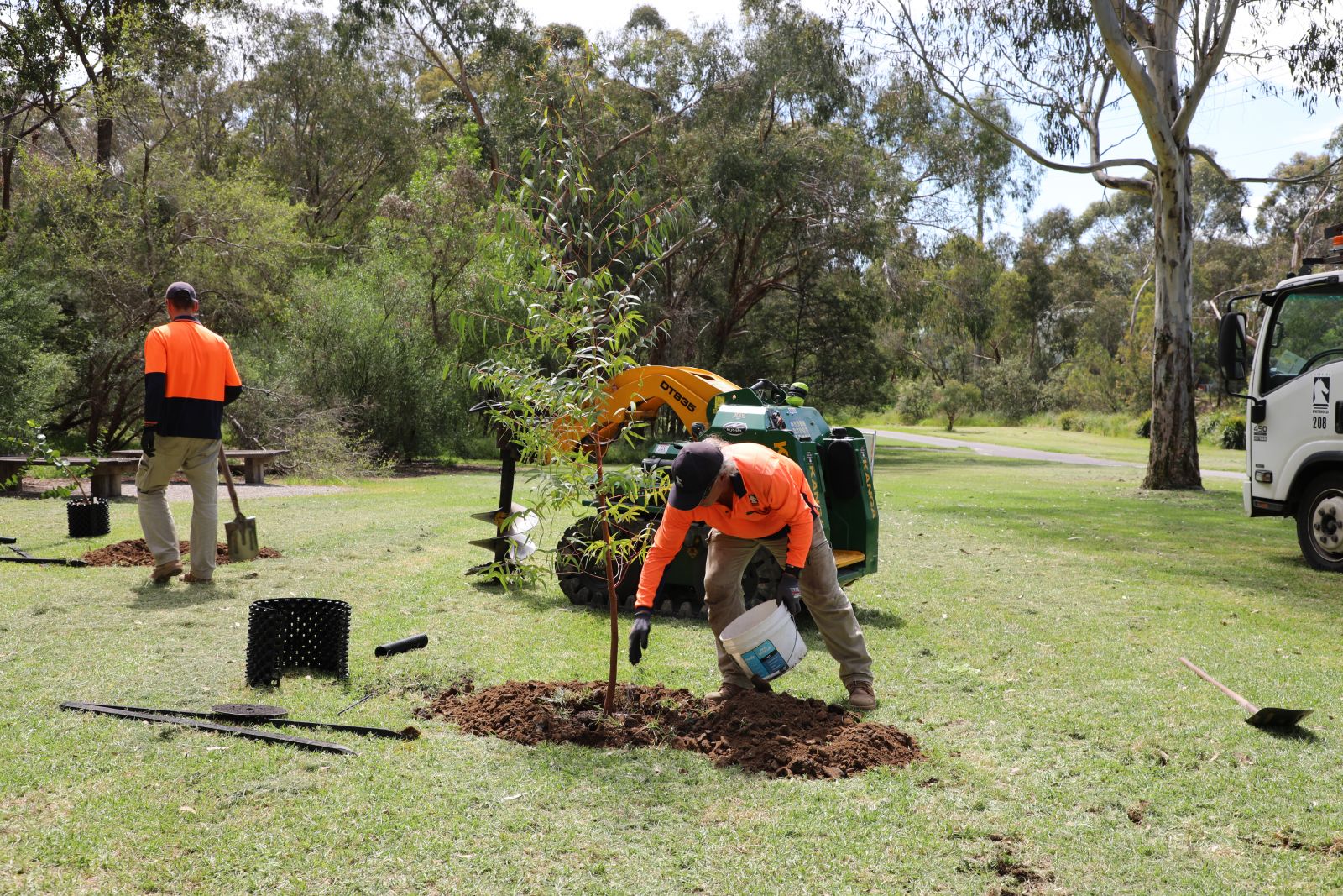 Two people planting trees in high vis, one holding a bucket and wearing gloves and one in the distance holding a shovel