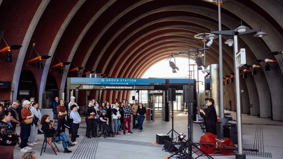 Audience members gather infront of artist Allara as she performs, with Arden Station, beneath the station's arches and wayfinding signage