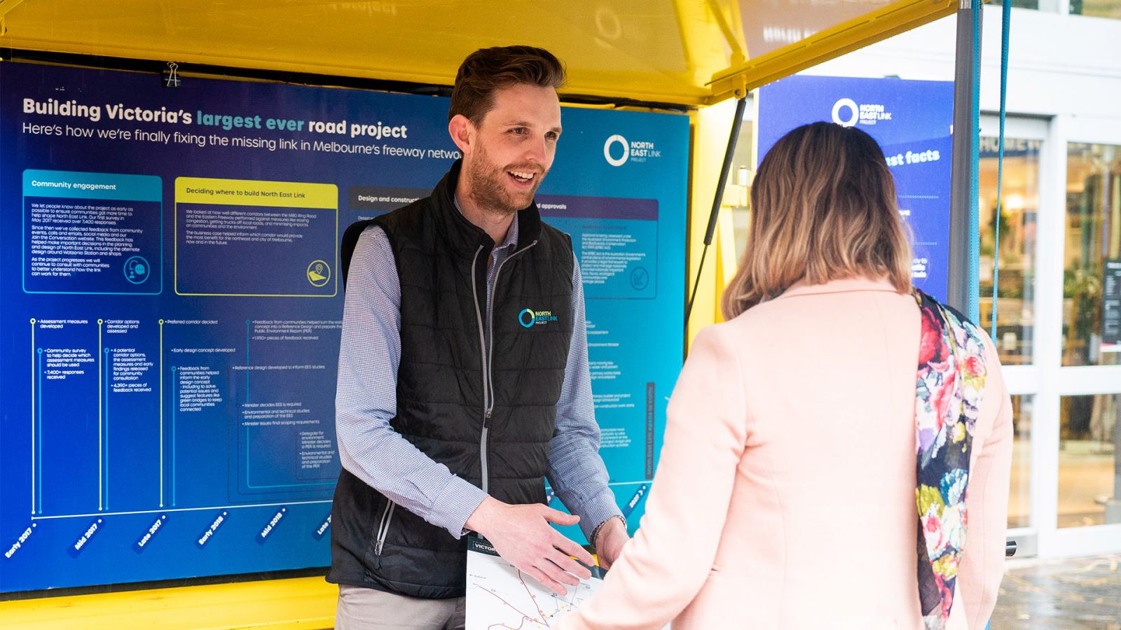 Two people talking to each other at an information session with North East Link graphics in the background.