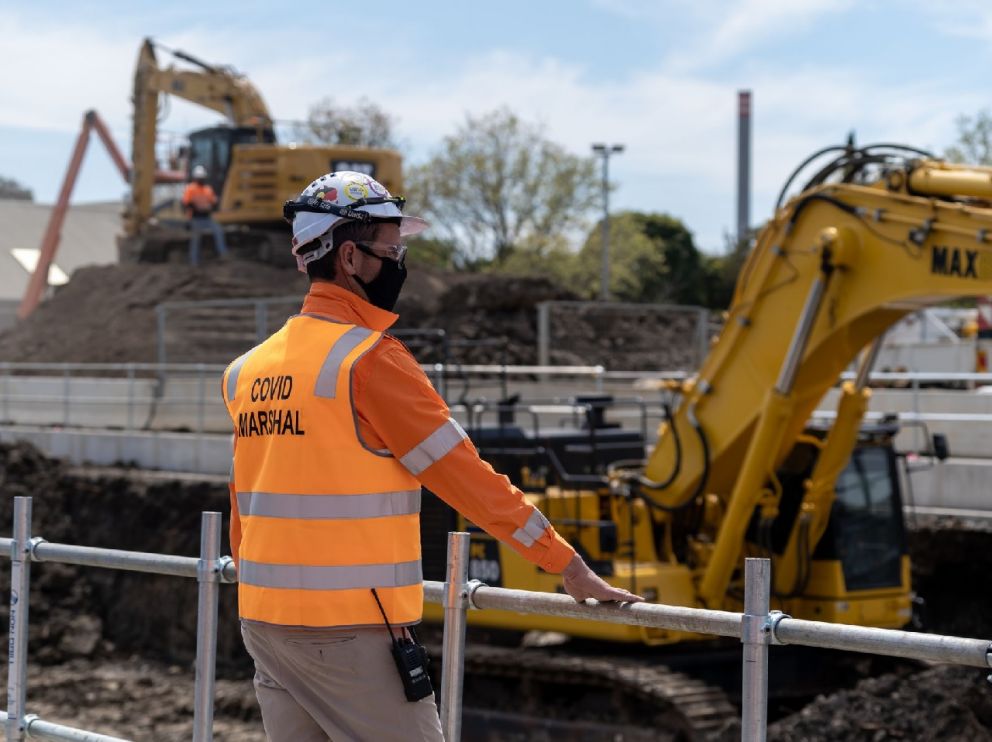 A man in an orange high-vis vest looks out over a worksite from behind fencing. His vest reads 'Covid Marshal' and he wears a mask and hard hat.