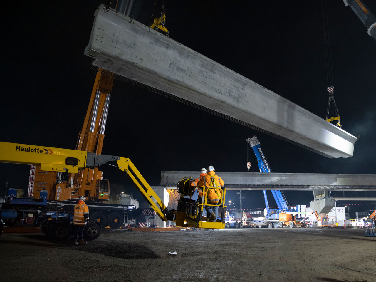 L-beam lifted in the air by a large crane with a construction site below.