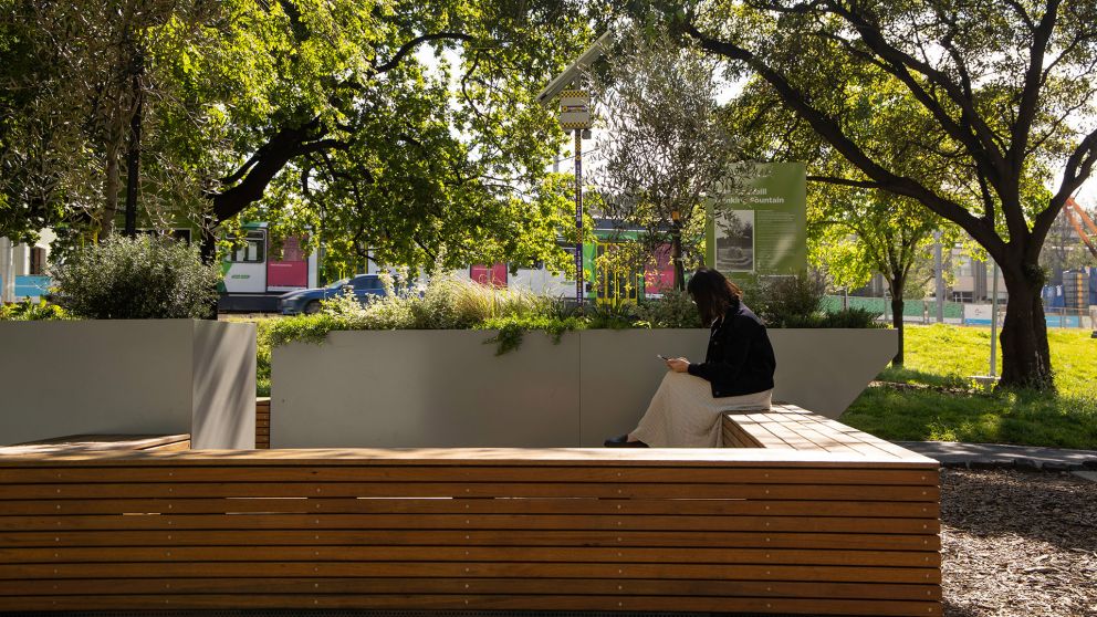 A person sits on a wooden bench which forms part of decking outdoors.