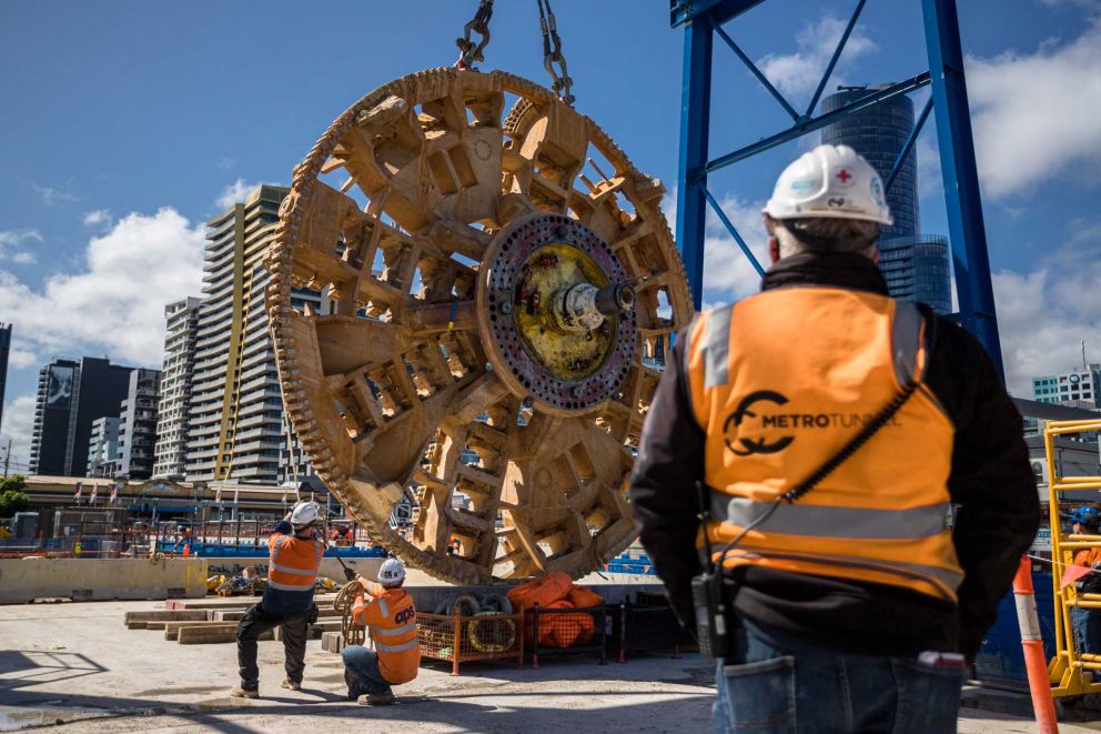 Workers in high vis clothing pull a cable attached to an enormous circular metal structure, hanging from a crane.