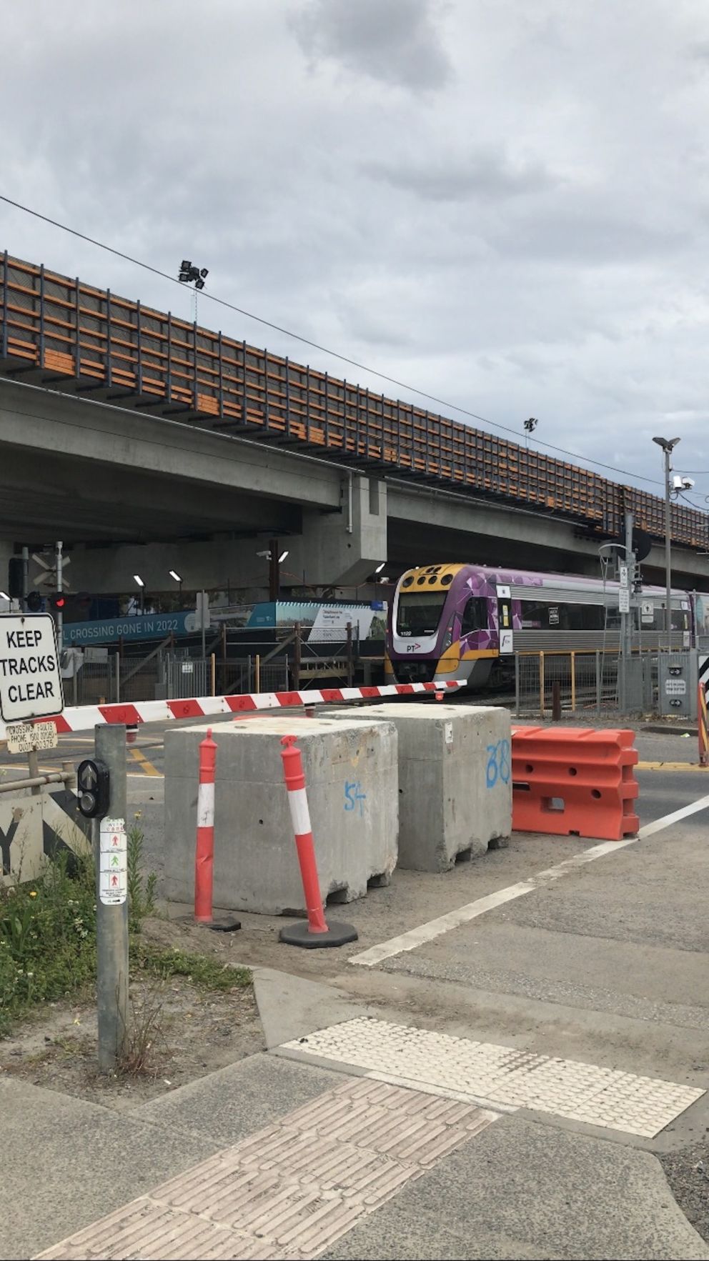 Boom gates down as a train passes through Hallam Road level crossing, with construction on the elevated rail bridge happening in the background.