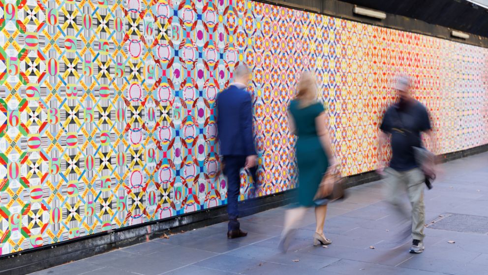 Three people walking past Elizabeth Gower's colourful artwork on a hoarding