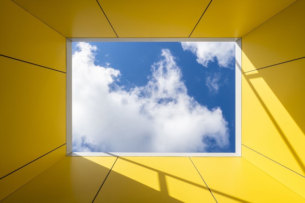 View looking up out of a skylight framed by yellow walls, with blue sky and clouds visible