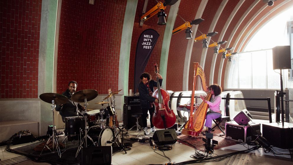 The Brandee Younger Trio performing beneath the arches at Arden Station, seated and playing the harp and various musical instruments with afternoon light shining in