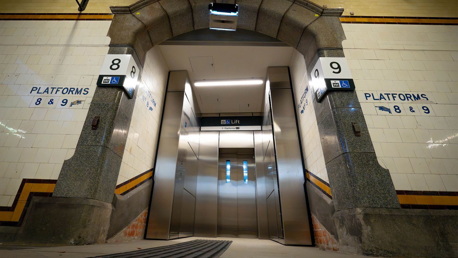 A lift flanked by signs indicating it connects with Platforms 8 and 9, located within a subway with decorative tiled walls.