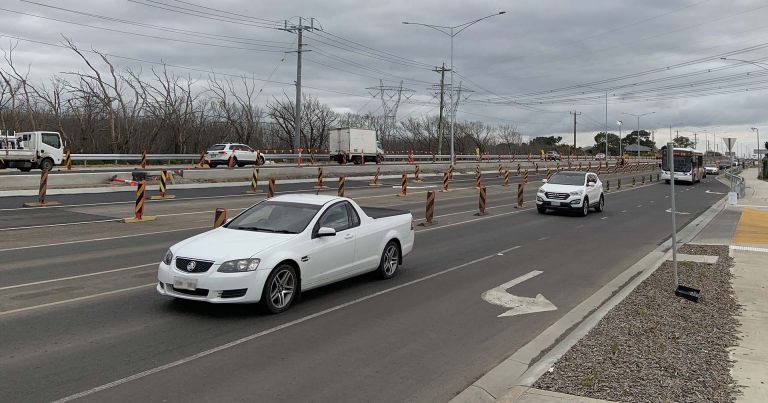 Newly constructed northbound lanes between Harvest Home Road and Craigieburn Road East, featuring active traffic with a white utility vehicle and SUV in the foreground. Orange and black striped barriers mark the construction zone, with trucks and other vehicles in the background. Power lines and poles line the road under an overcast sky.