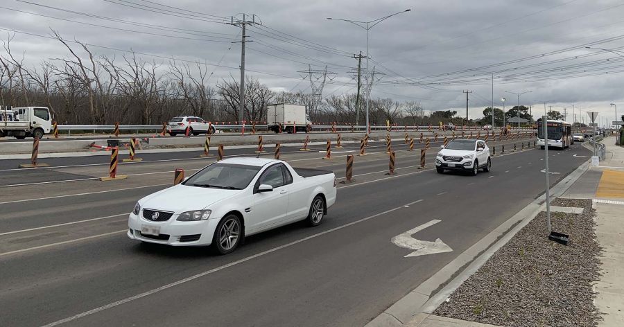 Newly constructed northbound lanes between Harvest Home Road and Craigieburn Road East, featuring active traffic with a white utility vehicle and SUV in the foreground. Orange and black striped barriers mark the construction zone, with trucks and other vehicles in the background. Power lines and poles line the road under an overcast sky.