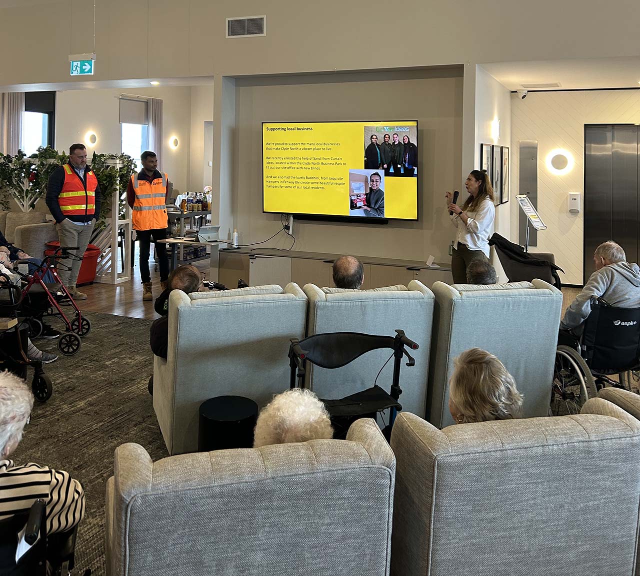 A group of people seated in a modern room watching a presentation on a large wall-mounted television screen. Three individuals in high-visibility vests stand to the left, while another person presents near the screen. Audience members include individuals using wheelchairs and walkers.