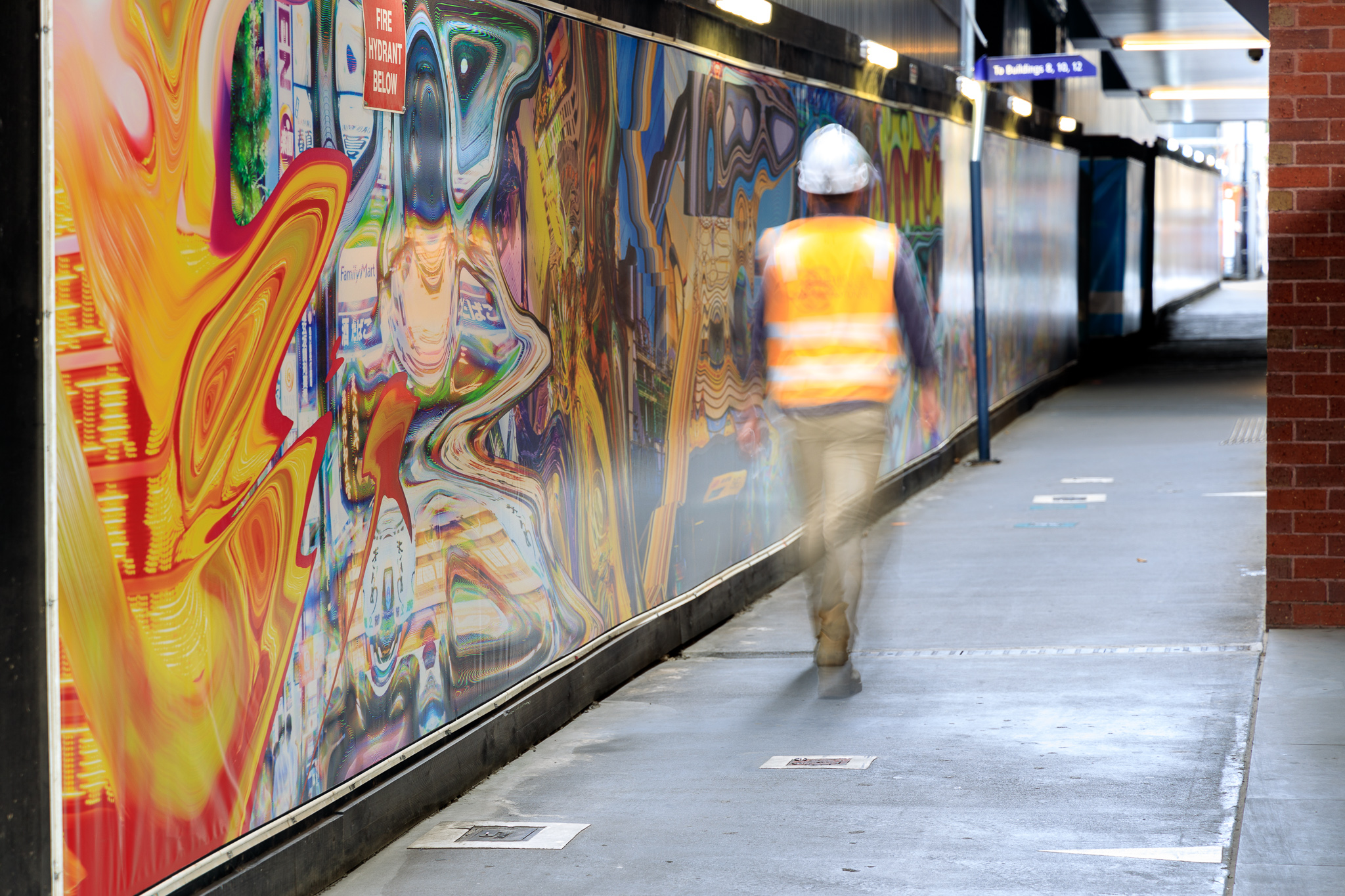 A person in a hard hat and hi-vis walks past a colourful and distorted image on a wall