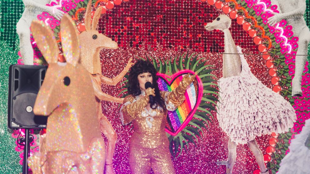 Woman dressed in a glitter body suit singing in front of the Huxley artwork