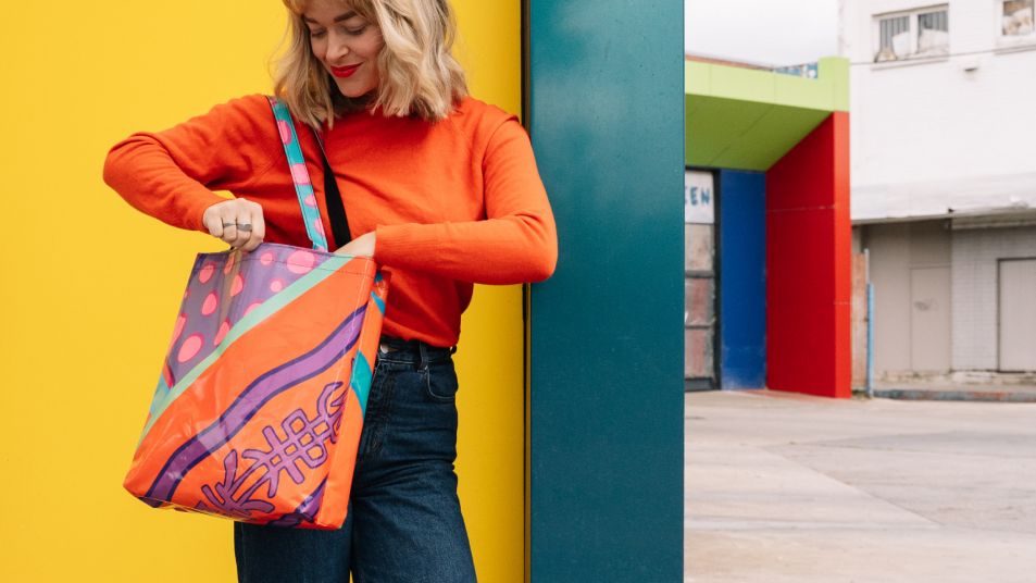 A person with long blonde hair wearing an orange coloured jumper is looking in their brightly coloured tote bag