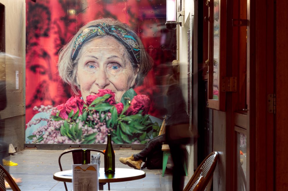 A bright artwork at the back of a bustling alleyway with a cafe, depicting an older woman's face on a pink background with flowers in the foreground