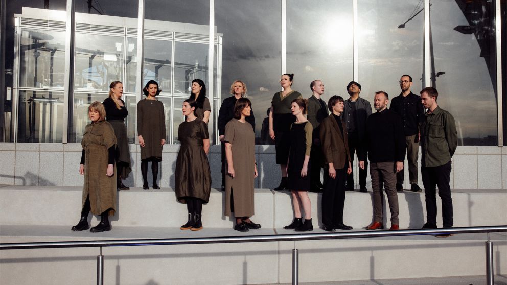 Vocal group Invenio, dressed in shades of khaki, black and brown, stand in two rows outside Arden Station facing different directions while they perform 