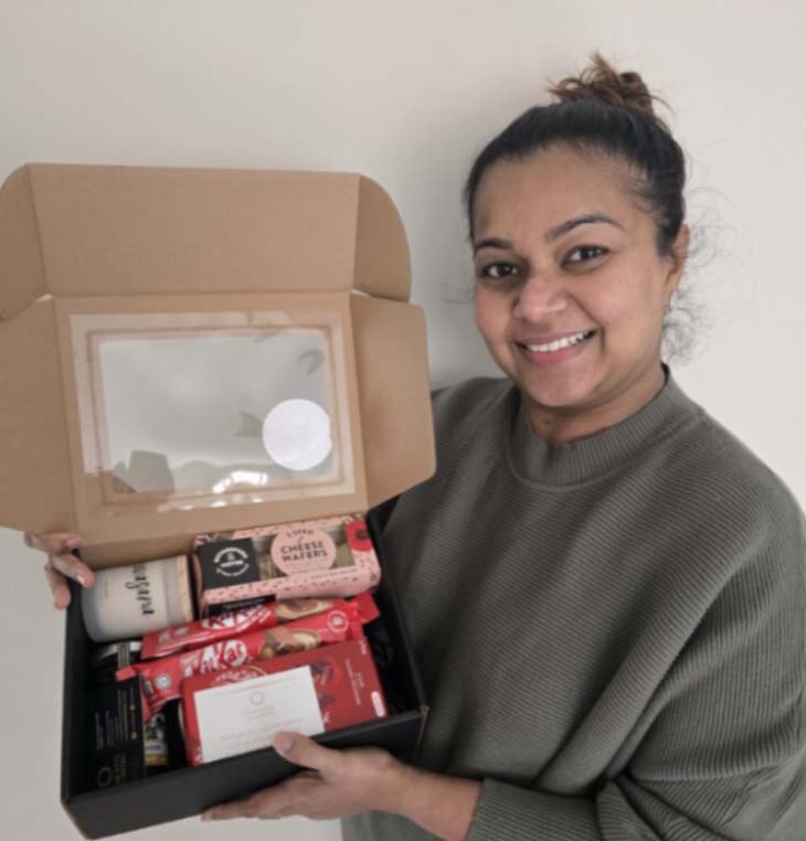 A person holding an open cardboard box containing assorted food and drink items. The woman is smiling and wearing a sweater, with a plain background behind them.